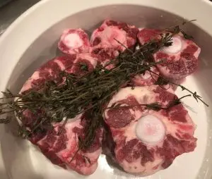 Washed and Sliced Oxtails in a white bowl with fresh springs of thyme for Single Ingredient Groceries.
